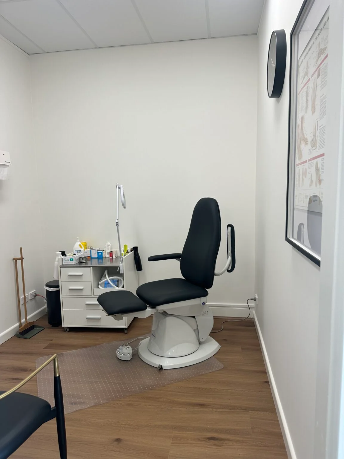 Clean podiatry clinic treatment room with a black examination chair and medical supplies.